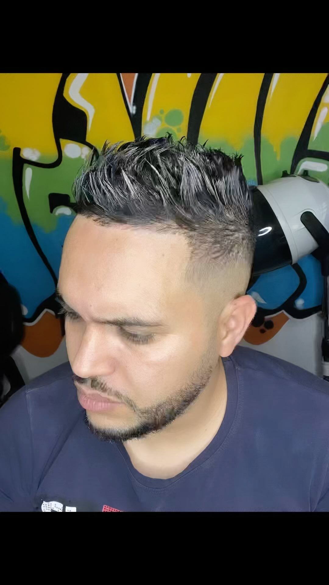 Hombre con corte de pelo descolorido y barba incipiente, mirando hacia abajo. Grafiti al fondo.