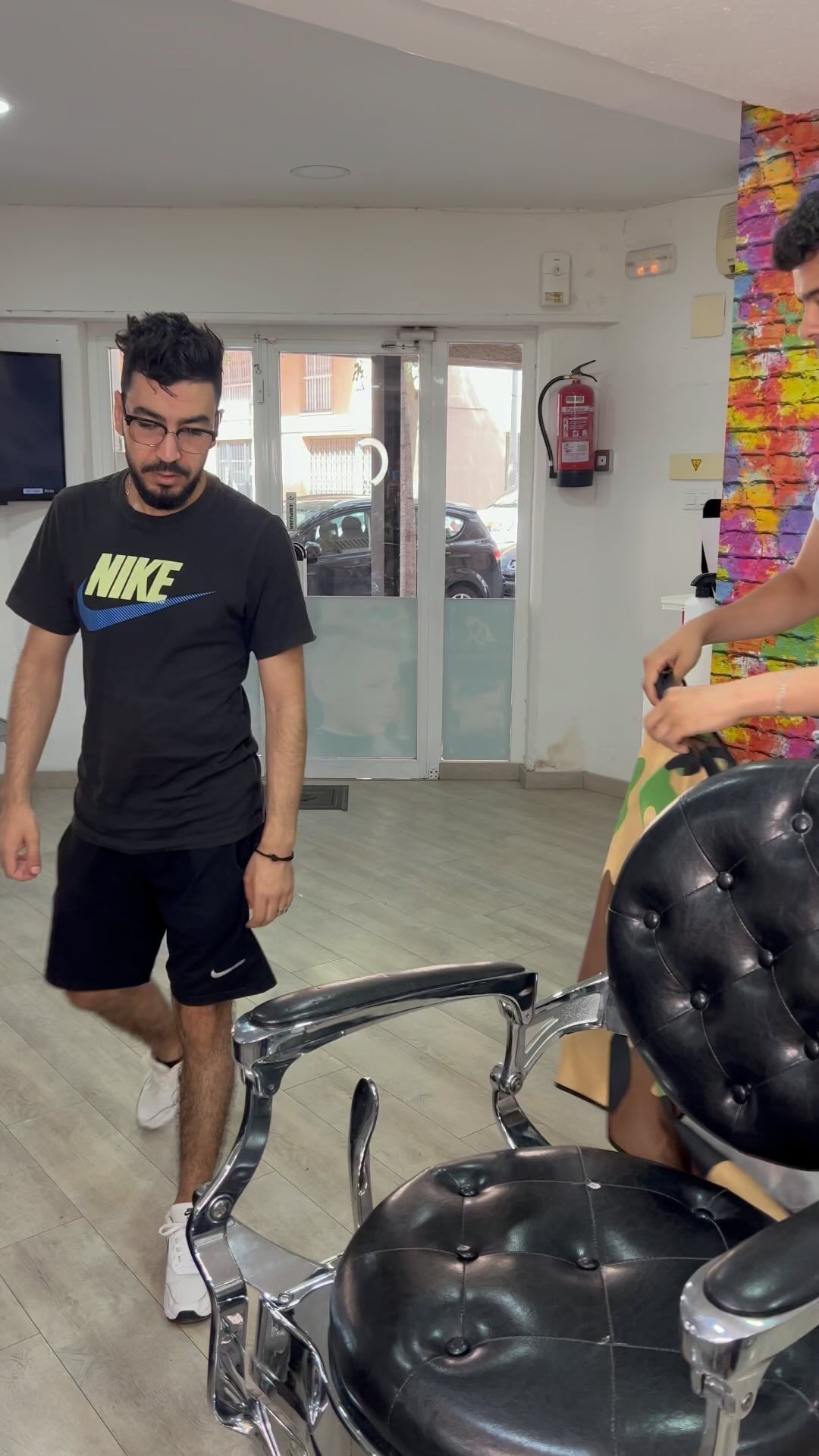 Un hombre camina en una barbería cerca de una silla de barbero. Otro hombre está de pie, con una maquinilla en la mano.