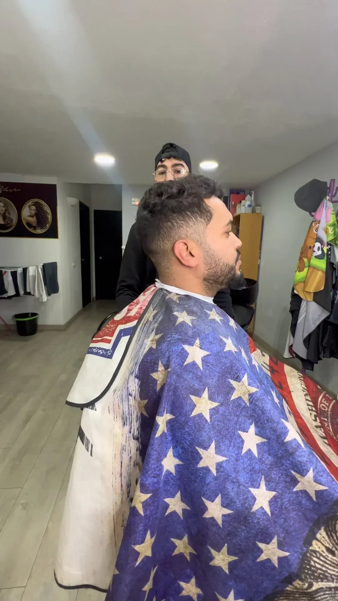 Un hombre se corta el pelo en una barbería, con una capa de estrellas. Un barbero está detrás de él.