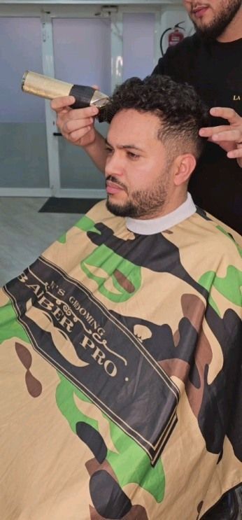Un hombre se corta el pelo en una barbería. El barbero usa una maquinilla. Lleva una capa de camuflaje.