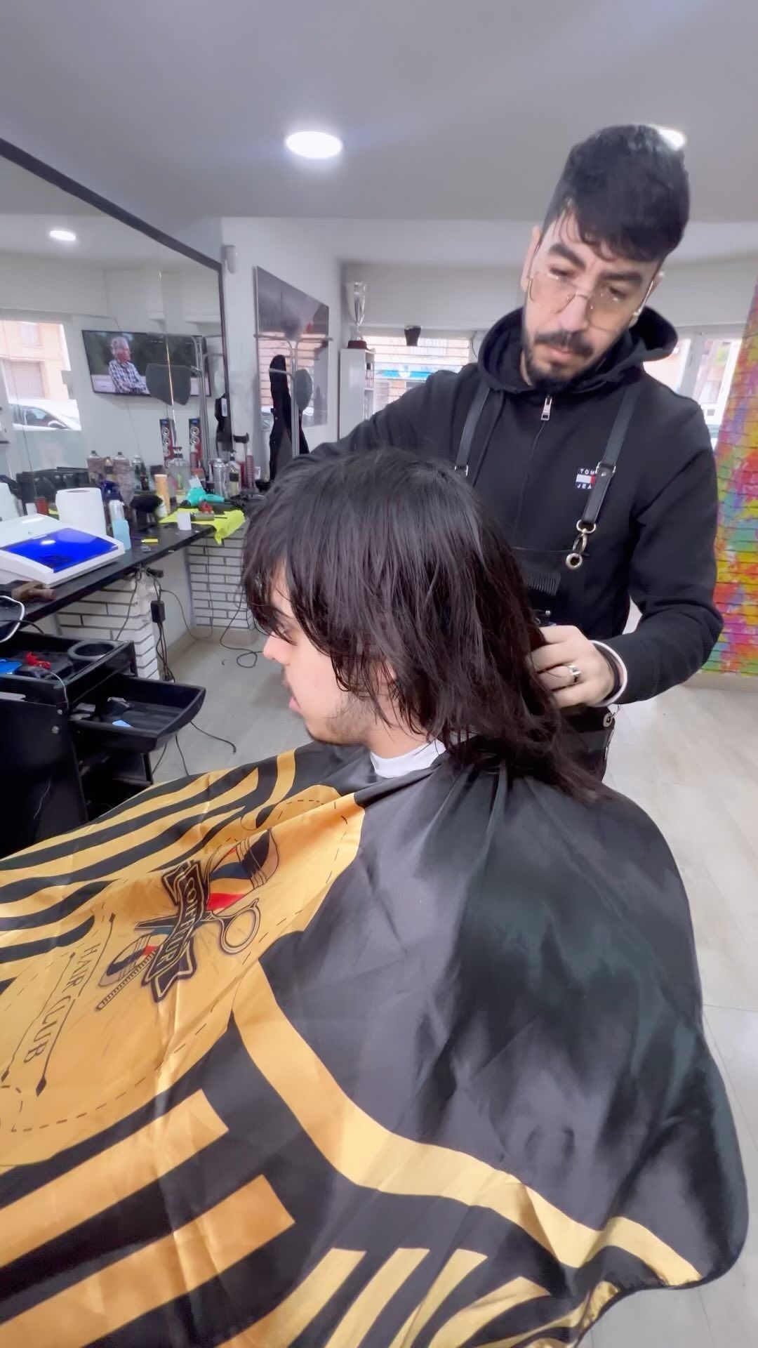 Un barbero corta el pelo mojado de una persona en un salón. La persona lleva una capa negra y dorada.