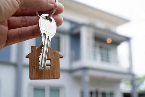 Una mano sostiene un llavero de madera con forma de casa, unido a una llave de metal, con una casa borrosa al fondo.