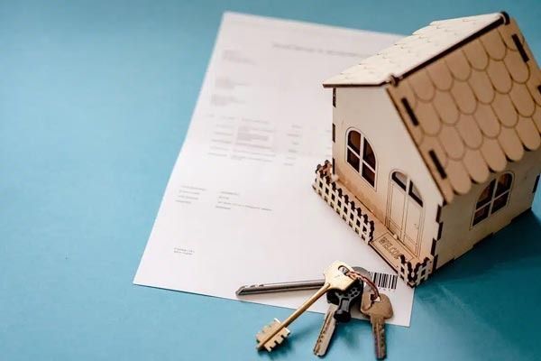 Sobre un documento, junto a un juego de llaves metálicas, se encuentra una pequeña maqueta de una casa de madera, sobre un fondo azul liso.