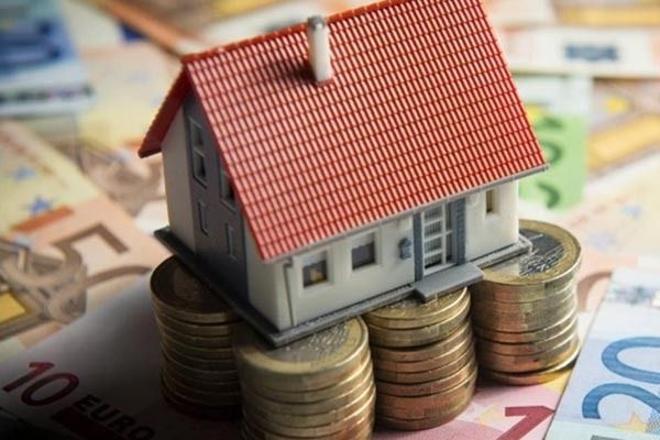Una maqueta de una casa con techo rojo se mantiene en equilibrio sobre tres pilas de monedas de oro, rodeada de varios billetes de euro.