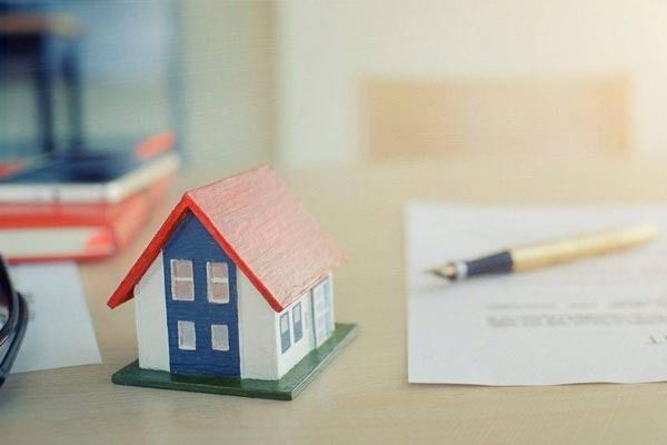 Una pequeña maqueta de una casa con techo rojo reposa sobre un escritorio junto a un documento y un bolígrafo.