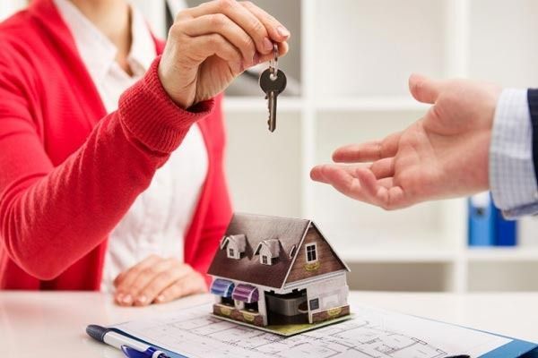 Una persona con un suéter rojo le entrega la llave de una casa a otra persona que está sobre un escritorio con una pequeña maqueta de una casa y un plano.