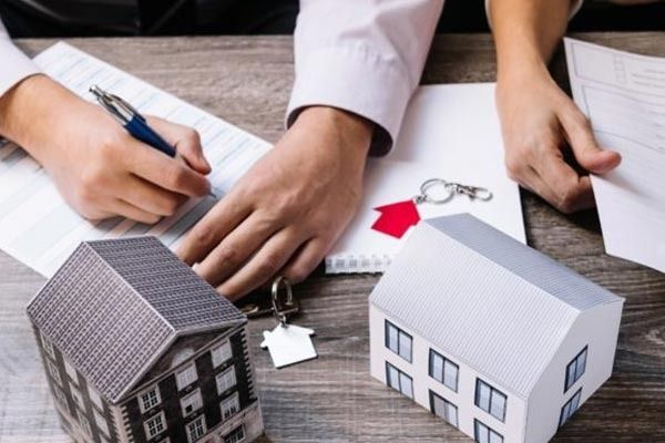Dos personas están sentadas a una mesa de madera firmando documentos inmobiliarios; sobre el escritorio hay dos pequeñas maquetas de casas y una llave.