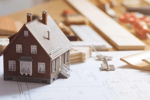Una maqueta de una casa reposa sobre planos arquitectónicos junto a una llave y herramientas de dibujo de madera.