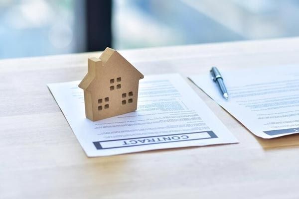 Sobre una mesa de madera de color claro, reposa una pequeña maqueta de una casa de madera, junto a un documento contractual, al lado de un bolígrafo.