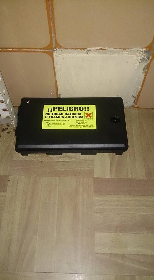 Una estación de cebo para roedores, de forma rectangular y negra, con una etiqueta de advertencia amarilla que dice 