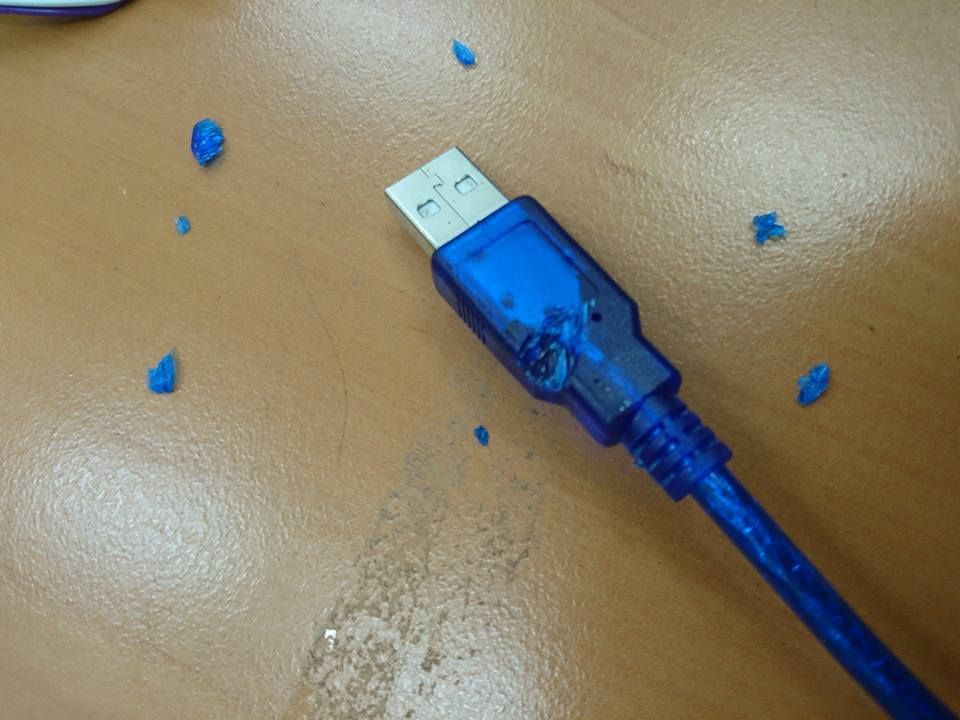 Un cable USB azul con una carcasa de conector dañada y derretida que reposa sobre una superficie de madera con pequeños fragmentos de plástico azul.
