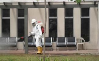 Una persona con equipo protector blanco y botas rocía desinfectante en sillas al aire libre frente a un edificio.