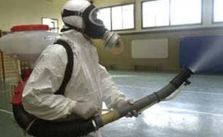 Una persona con un traje de protección que cubre todo el cuerpo y una máscara de gas utiliza un pulverizador de mochila para desinfectar un gimnasio.