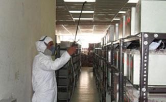 Una persona con equipo de protección blanco y una máscara utiliza un pulverizador de largo alcance para desinfectar los estantes de un almacén.