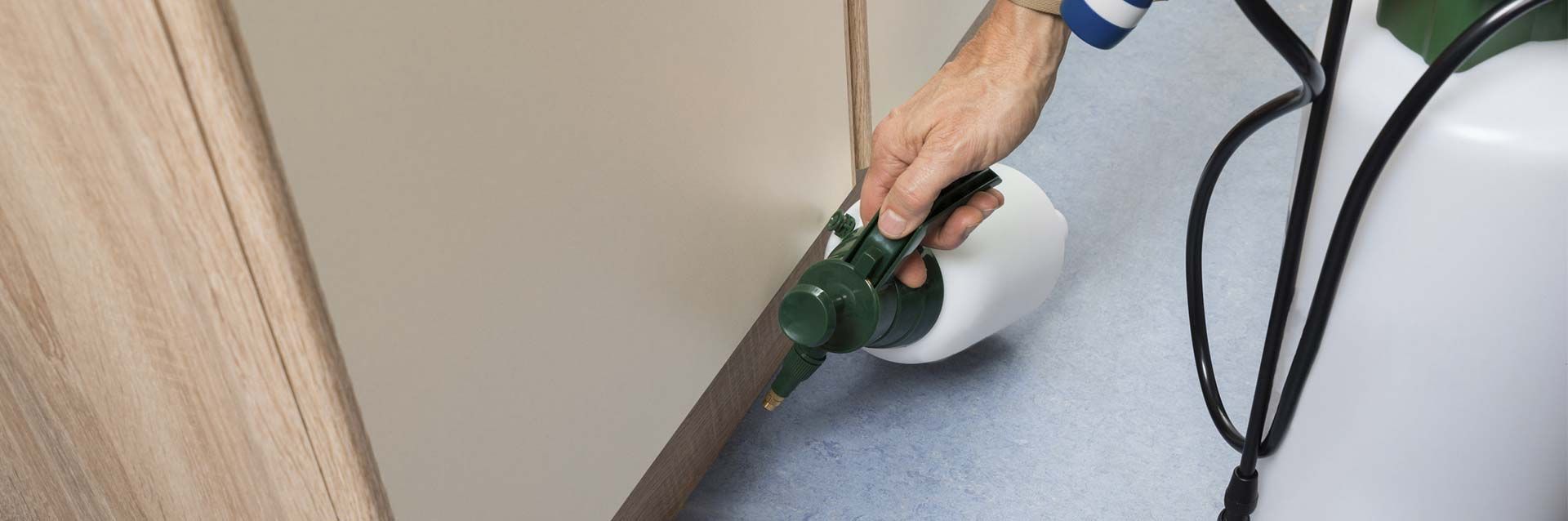 Una mano utiliza un pulverizador manual verde y blanco para aplicar un tratamiento a lo largo del borde de una pared y el suelo.