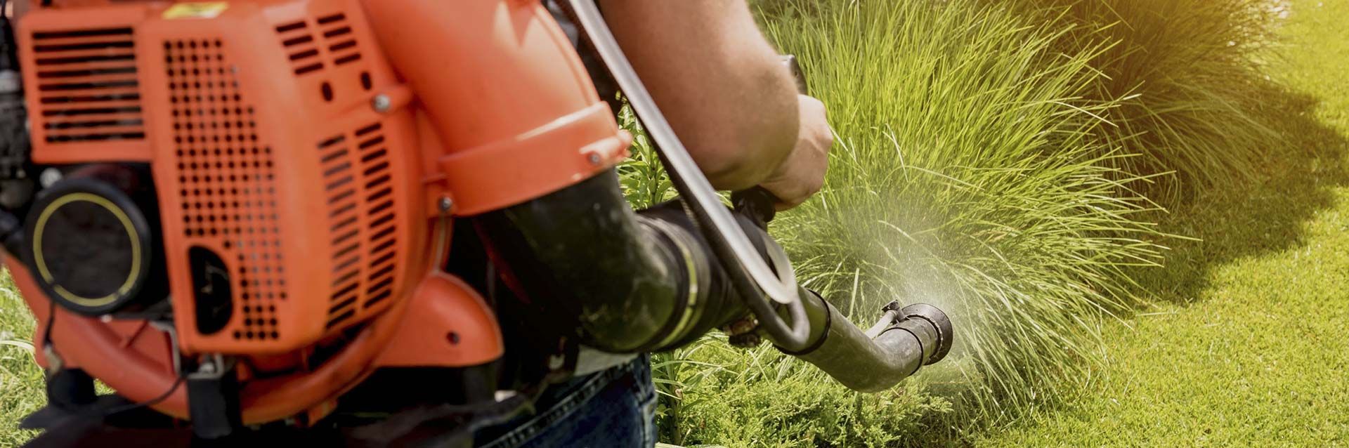 Una persona opera un soplador de hojas con mochila de color naranja para limpiar los recortes de césped de un césped.