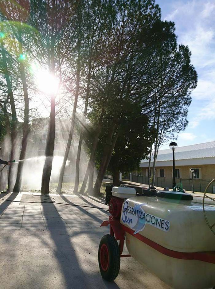 Una unidad pulverizadora móvil en un parque iluminado por el sol, rociando niebla hacia una hilera de árboles cerca de un edificio.