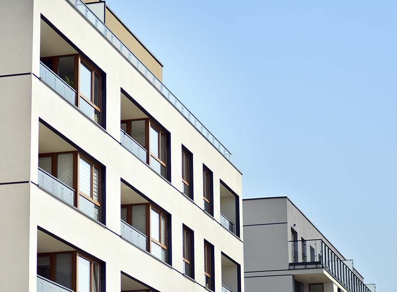 Modernos edificios de apartamentos con fachadas blancas, balcones con barandillas de vidrio y grandes ventanas contra un cielo azul claro.