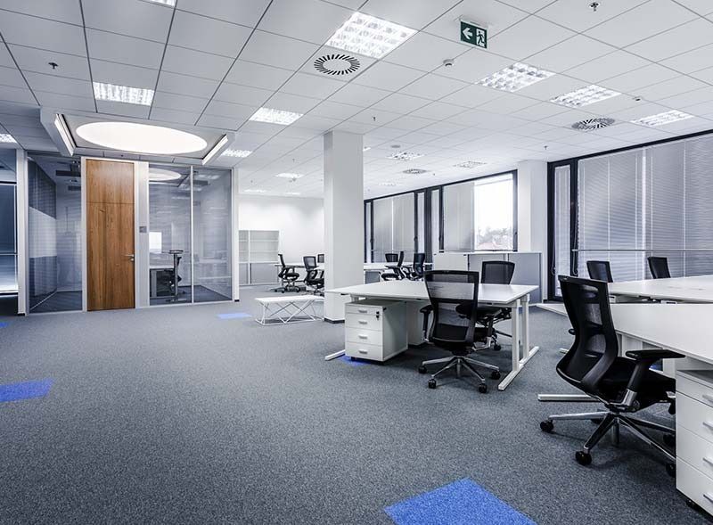 Una oficina abierta con varios escritorios, sillas ergonómicas negras, alfombra gris y una gran puerta de madera.