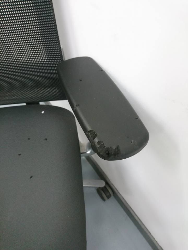 Una silla de oficina negra con un apoyabrazos dañado y roto y varios agujeros pequeños en el cojín del asiento.