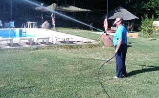 Una persona con una camisa azul rocía un chorro de agua con una manguera cerca de una piscina al aire libre en un día soleado.