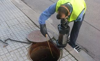 Un trabajador de servicios públicos con un chaleco de alta visibilidad utiliza una bomba para dar servicio a un pozo de acceso abierto en la calle.