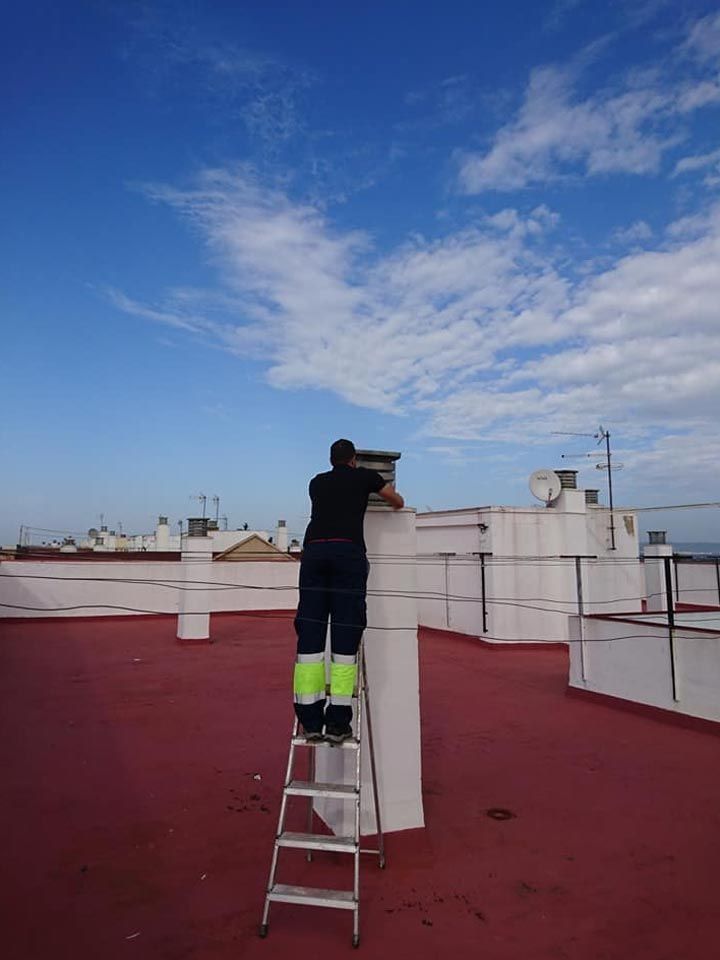 Una persona con ropa de trabajo oscura y bandas de alta visibilidad en las piernas se encuentra de pie en una escalera de mano trabajando en la chimenea de un techo.