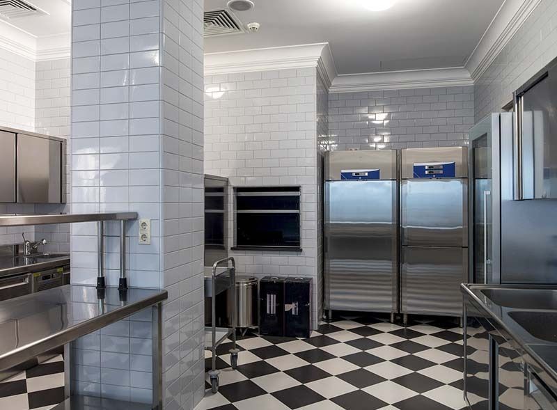 Cocina comercial con electrodomésticos de acero inoxidable, paredes de azulejos blancos tipo metro y piso a cuadros en blanco y negro.