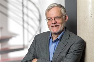 Ein Mann in Anzug und blauem Hemd lehnt mit verschränkten Armen an einer Wand.