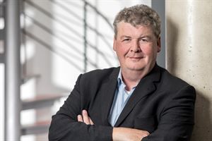 Ein Mann im Anzug lehnt mit verschränkten Armen an einer Wand.