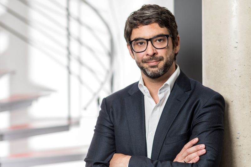 Ein Mann in Anzug und Brille lehnt mit verschränkten Armen an einer Wand.