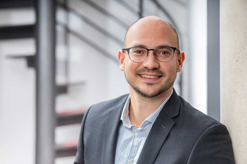 Ein Mann im Anzug und mit Brille lehnt an einer Wand und lächelt.