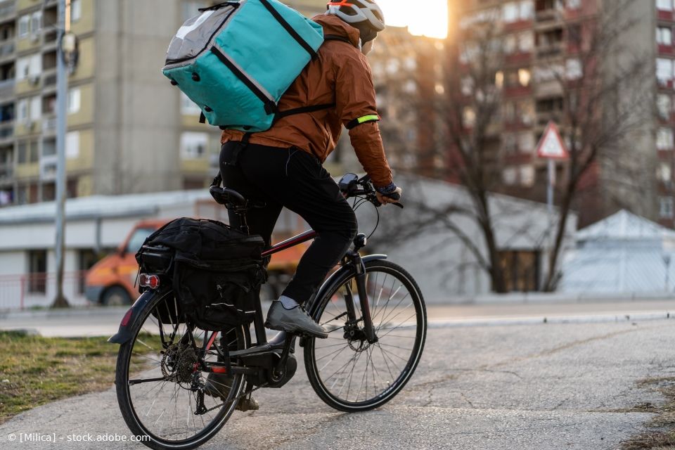 Eine Person mit einer Tasche für Essenslieferungen auf dem Rücken fährt auf einem Fahrrad auf einer Straße in der Stadt.