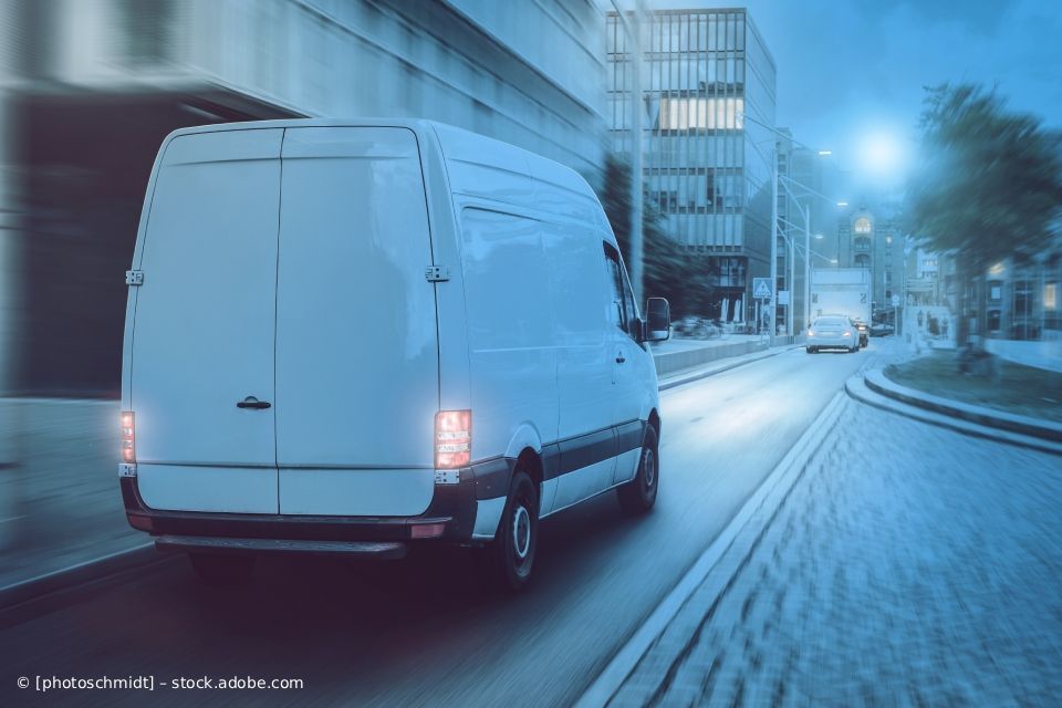 Ein weißer Lieferwagen fährt nachts durch eine Stadtstraße; im Hintergrund sind Gebäude und ein weiteres Fahrzeug zu sehen.
