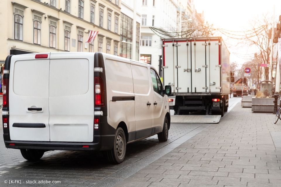 Ein weißer Lieferwagen und ein LKW parken auf einer Kopfsteinpflasterstraße; im Hintergrund sind Gebäude zu sehen.