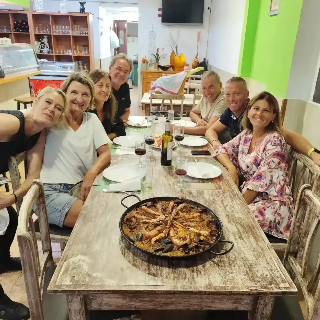 Un grupo de amigos alrededor de una mesa con un plato de paella, sonriendo en un entorno de restaurante.