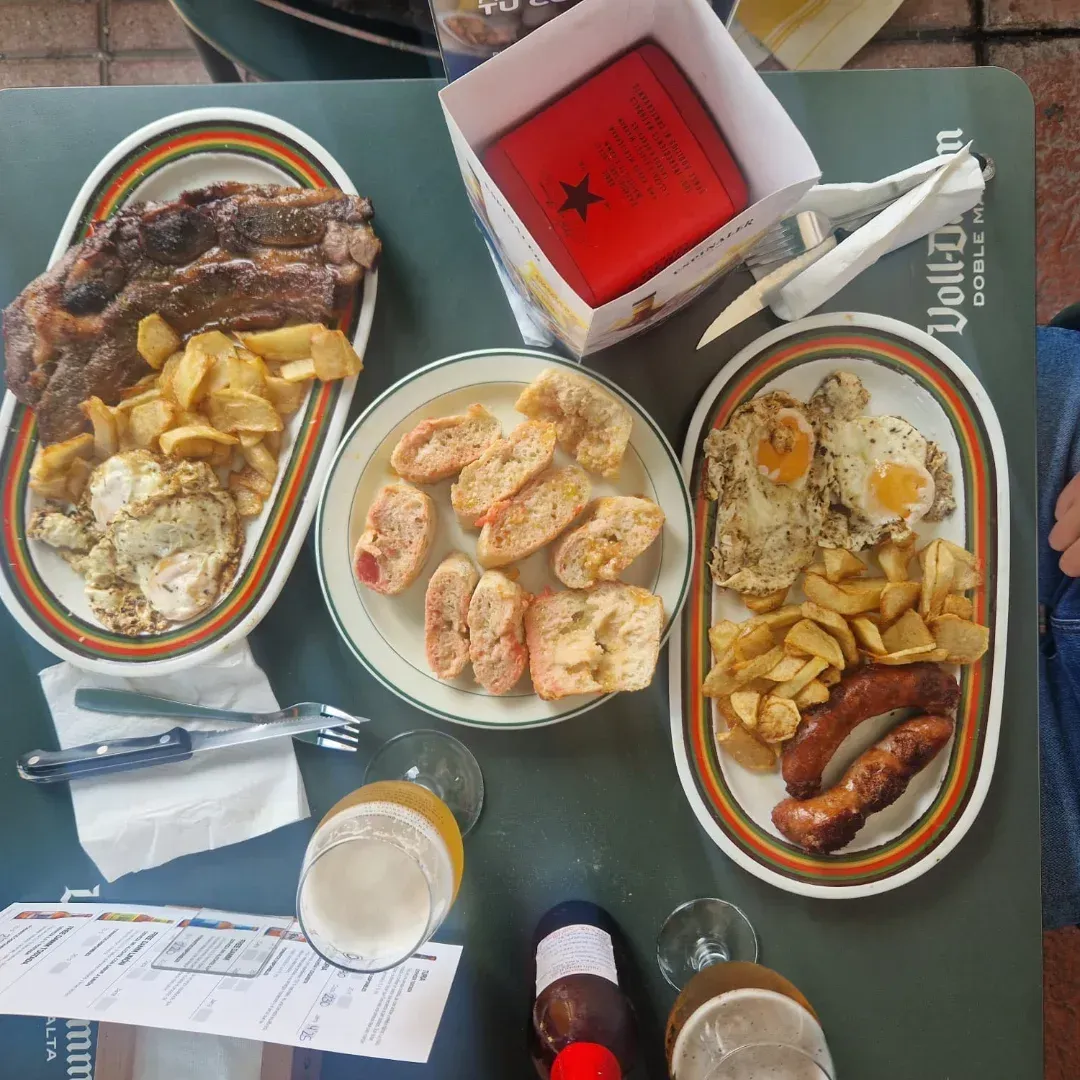 Fotografía cenital de una mesa con platos de comida, vasos de cerveza y una pequeña caja; bistec, salchichas, huevos y papas fritas.