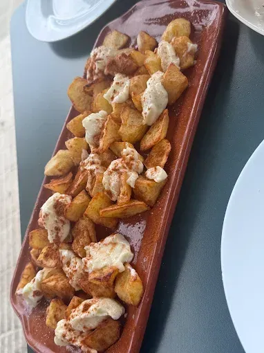 Patatas bravas en fuente rectangular de terracota, coronadas con alioli y pimentón, sobre una mesa oscura.