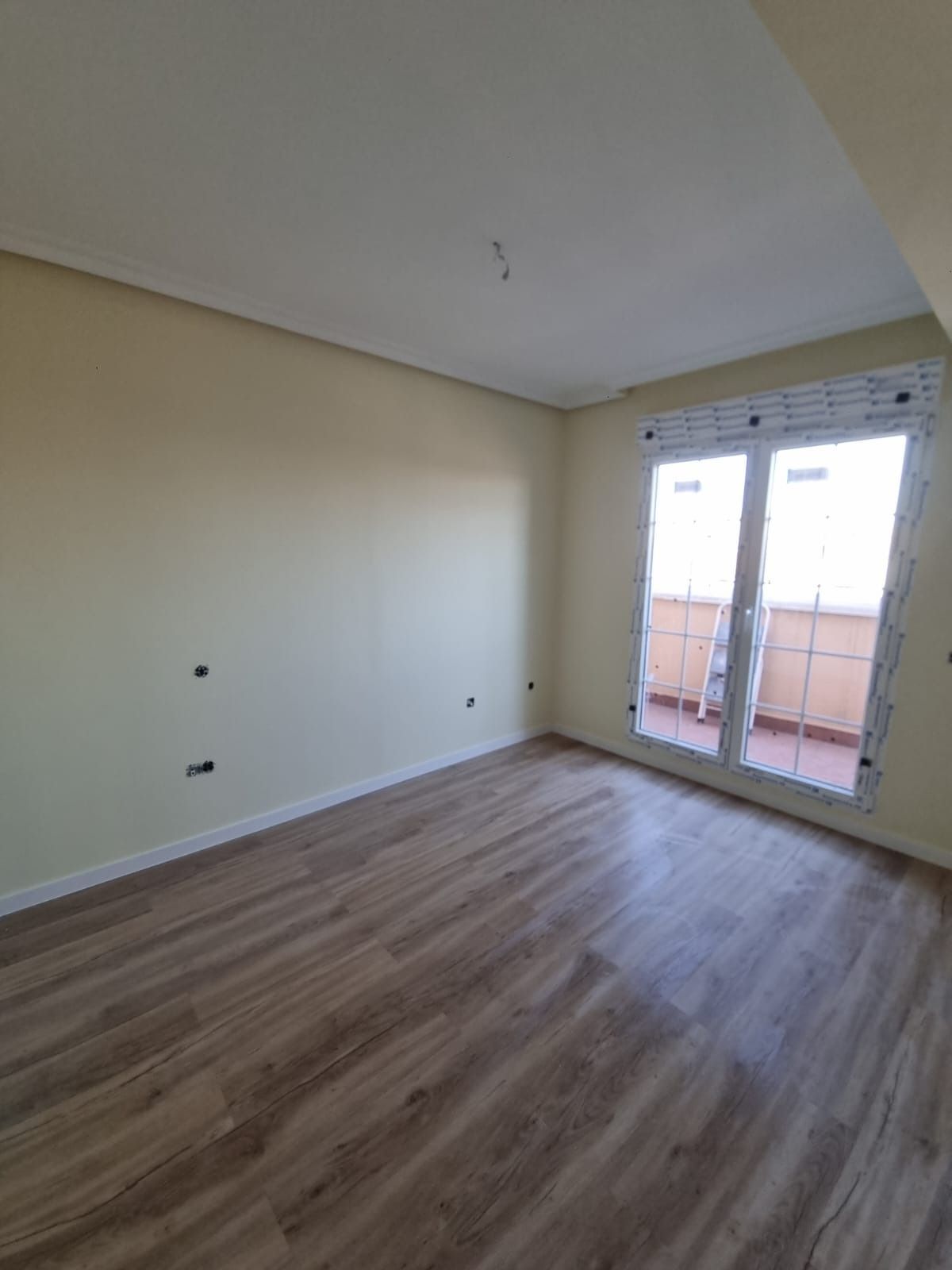 Habitación vacía con piso de apariencia de madera, paredes de color crema y una puerta de balcón con molduras blancas.