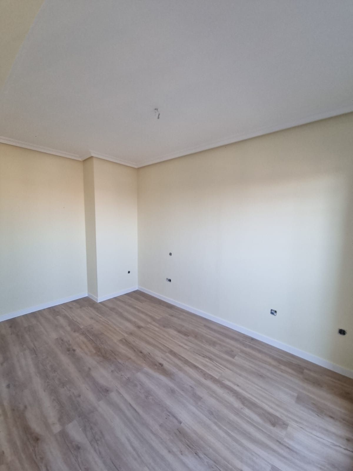 Habitación vacía con paredes beige, techo blanco y suelo imitación madera.