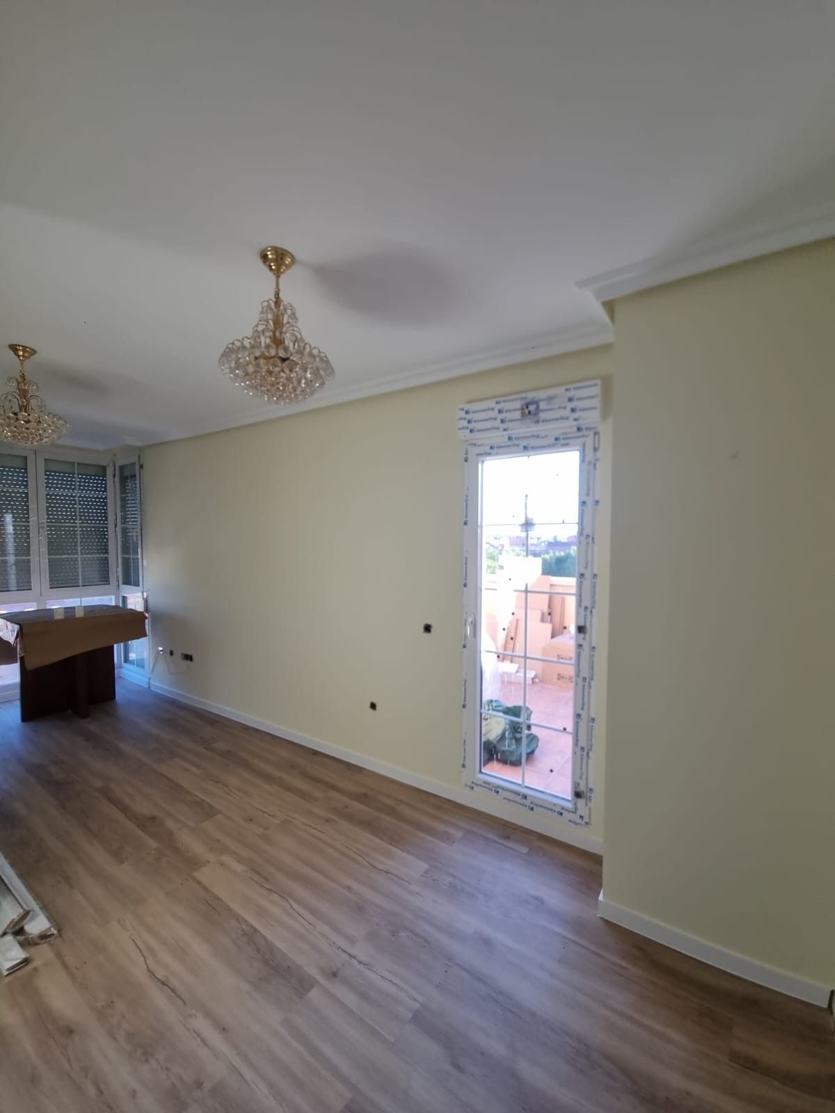 Sala de estar vacía con paredes de colores claros, pisos de madera, una puerta y dos candelabros de cristal.