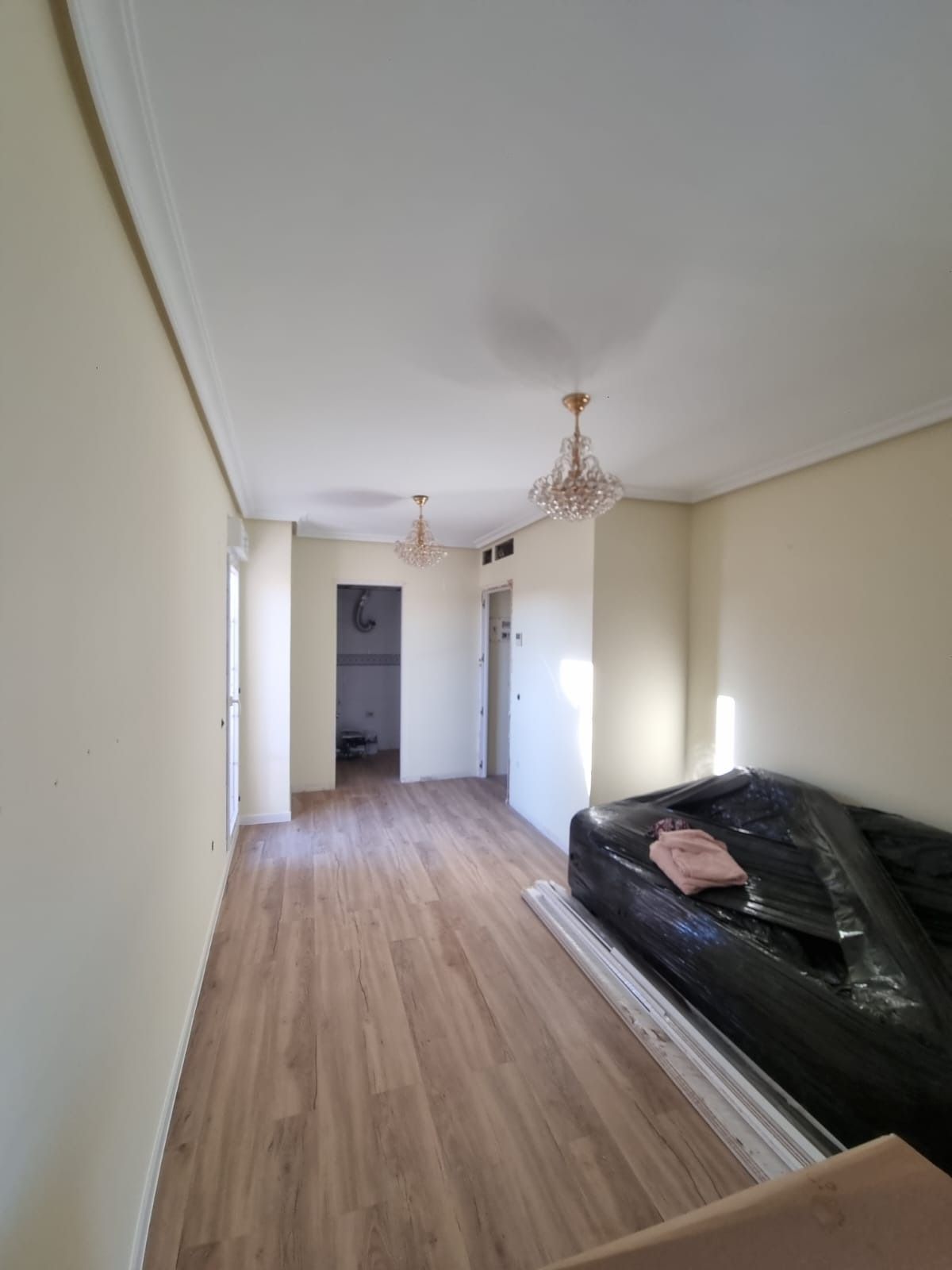 Habitación vacía con suelos de madera, paredes beige, molduras de corona y lámparas de araña. Un sofá tapizado a la derecha.