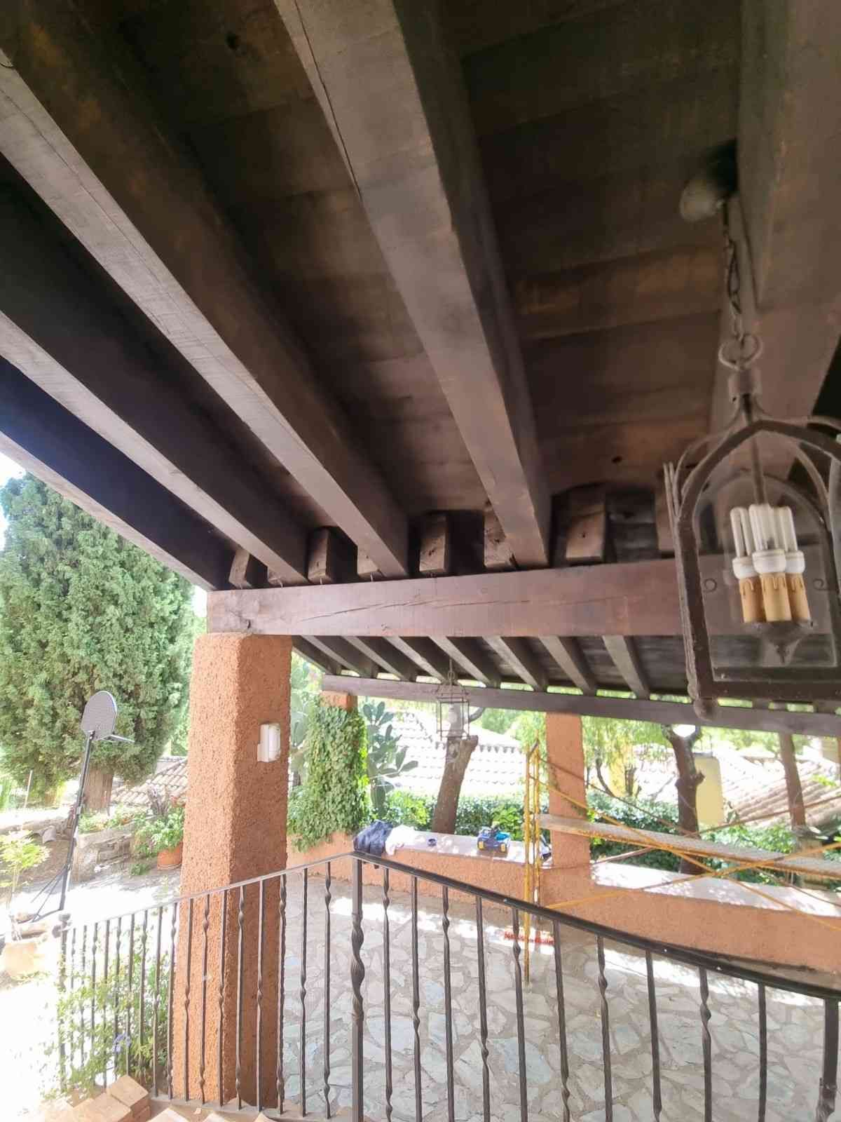 Vigas de madera que sostienen un techo marrón sobre un patio exterior con barandilla de metal y escalones de piedra.