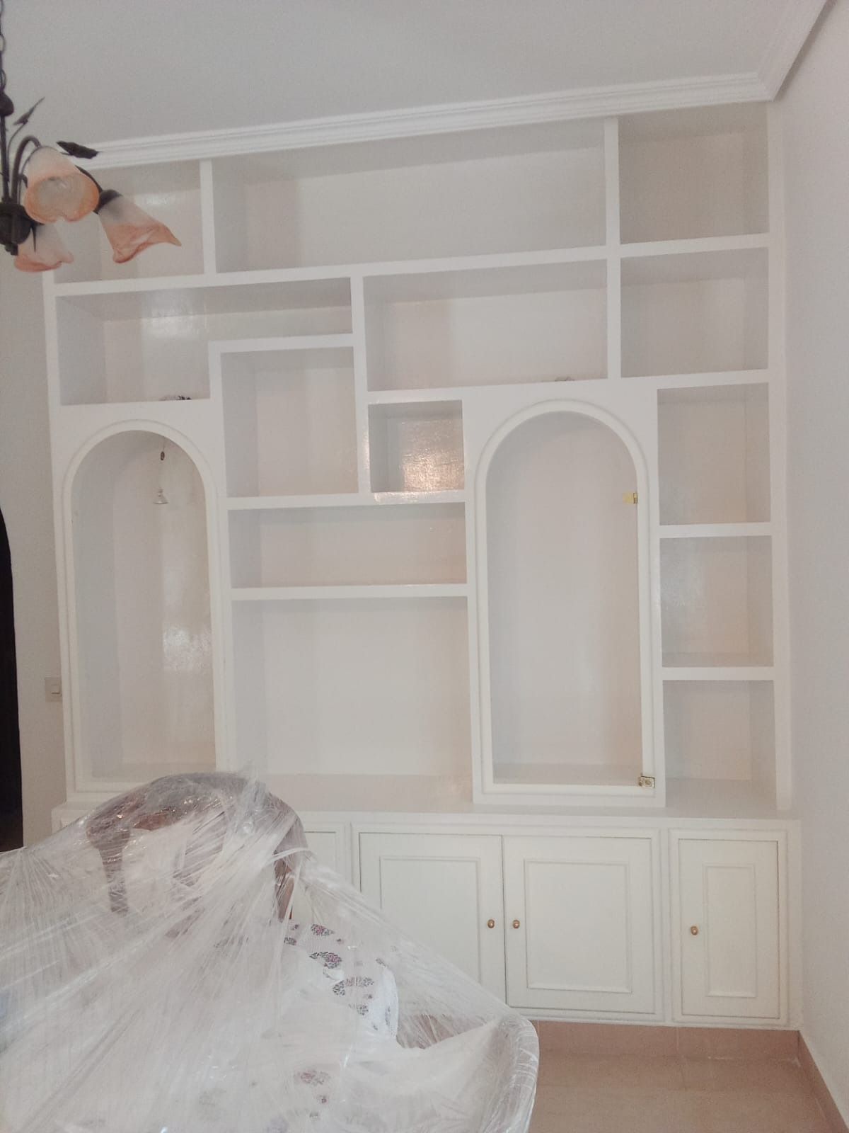 Estantería empotrada de color blanco con compartimentos de varios tamaños y nichos arqueados, sobre puertas de armarios.