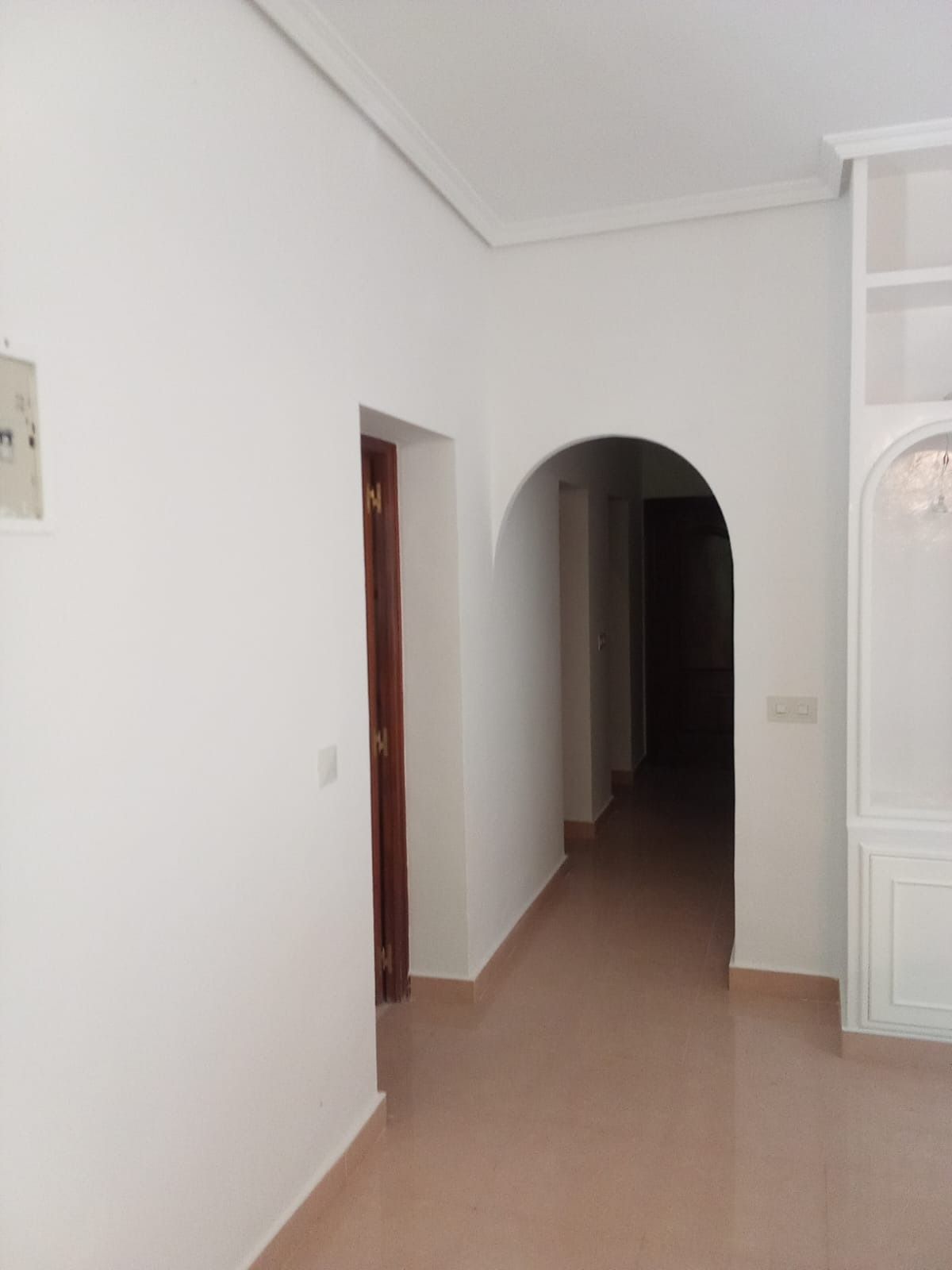 Pasillo blanco con una puerta arqueada que da a un pasillo más oscuro. Se ve una puerta de madera a la izquierda.