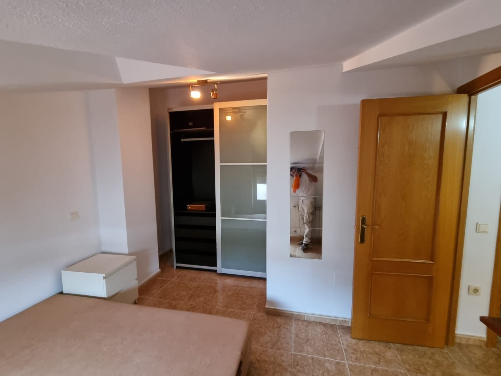 Dormitorio con armario, cama, suelo de baldosas marrones, puerta de madera y espejo de cuerpo entero.