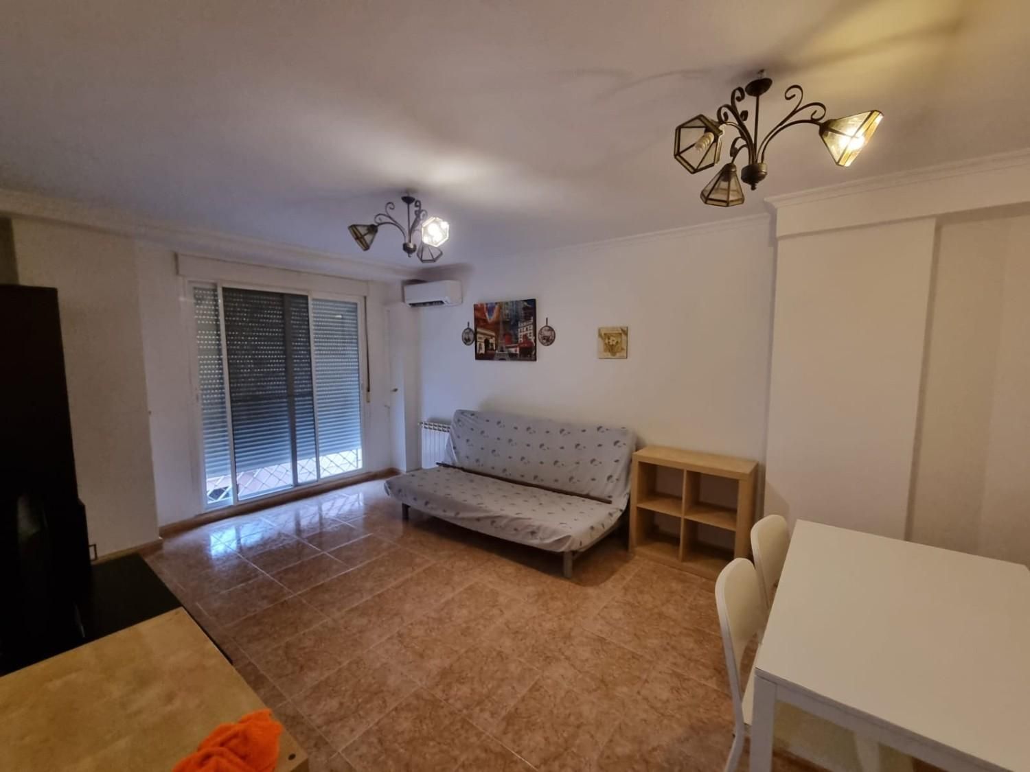 Sala de estar con sofá, mesa y puerta corredera. Suelo de baldosas marrones, paredes blancas y dos lámparas de araña.