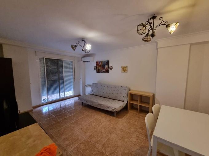 Sala de estar con sofá, mesa y puerta corredera. Suelo de baldosas marrones, paredes blancas y dos lámparas de araña.
