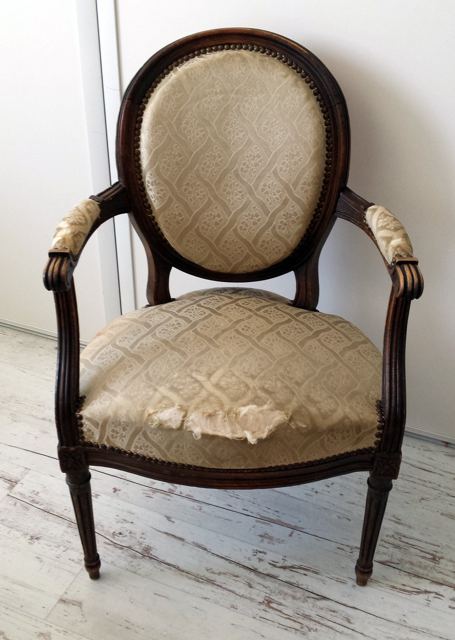 Fauteuil ancien beige capitonné, avec structure en bois foncé et coussin d'assise usé, adossé à un mur blanc