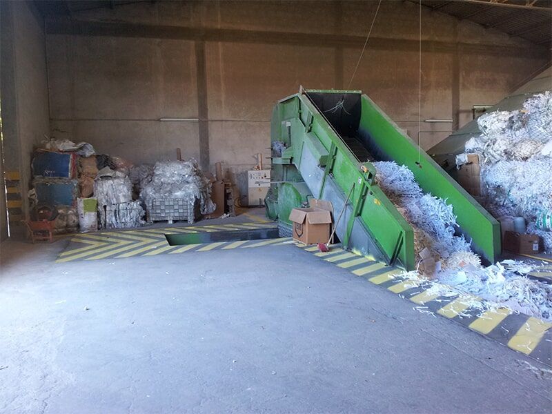 Uma máquina industrial verde de reciclagem de papel em um armazém. Pilhas de papel picado e caixas de papelão são visíveis.